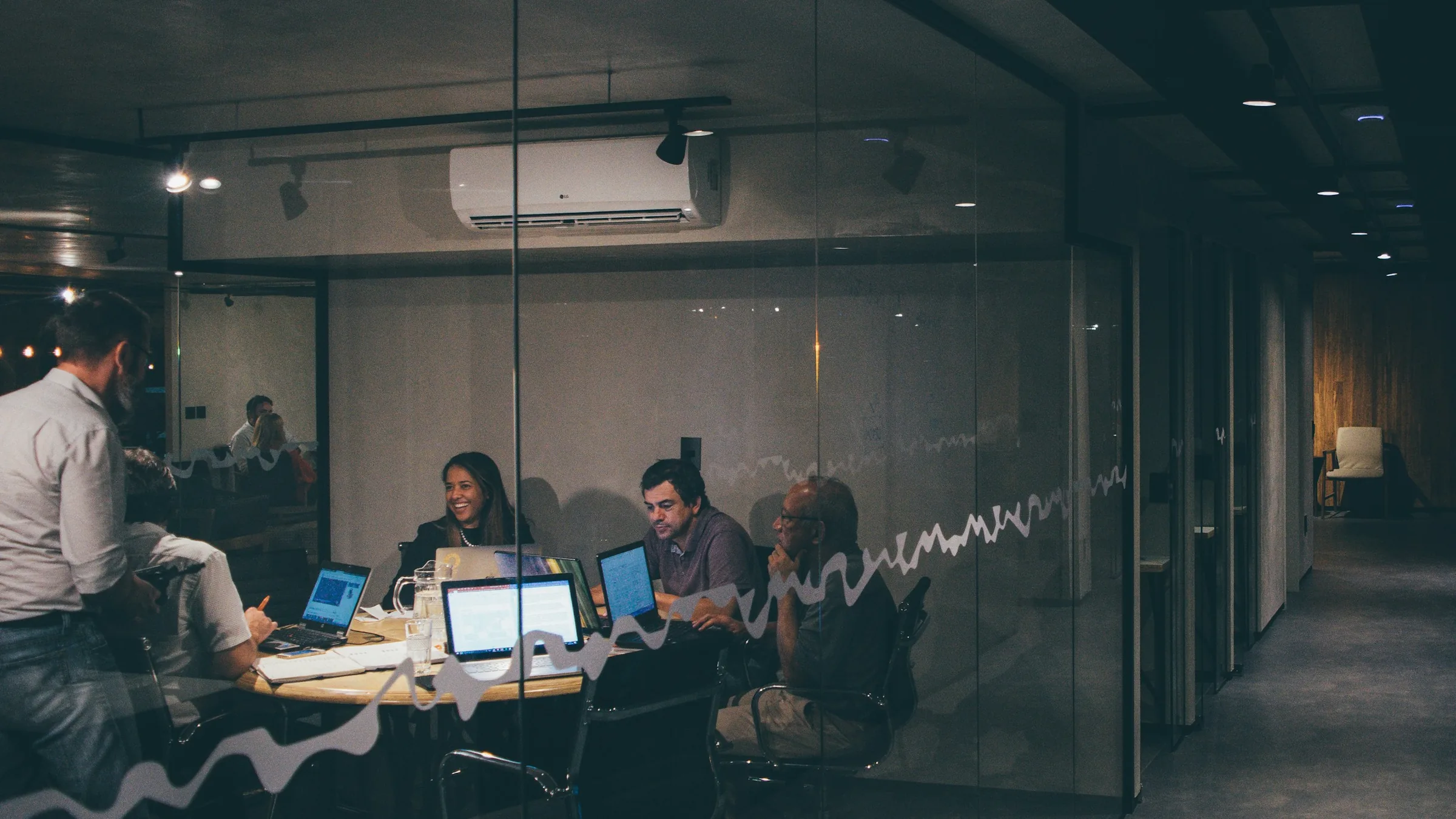 A small group working around one table late in the evening, seen through glass from a dark corridor.